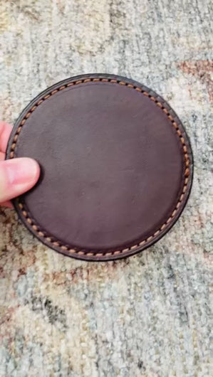 Video of a thick brown leather round coaster showing the top and bottom of the coaster and then showing a glass being placed on top of the well made coaster