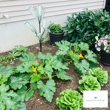 Load image into Gallery viewer, A full garden with the bottle tree yard art pictured in the middle of various green plants.