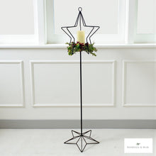 Load image into Gallery viewer, Decorative star-shaped candle holder with a candle and greenery against a white paneled wall.