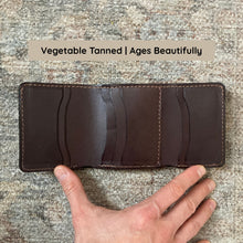 Load image into Gallery viewer, Brown leather wallet held by a hand on a textured surface with text 'Vegetable Tanned | Ages Beautifully'.
