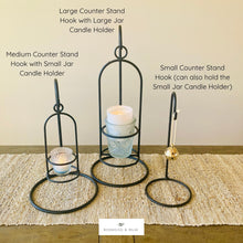Load image into Gallery viewer, Three candle holders on a wooden surface with labels indicating their size and use. The left and middle wrought iron tabletop hook stands are holding a glass jar candle.  The right and smaller display hook stand is holding a gold bell Christmas ornament.
